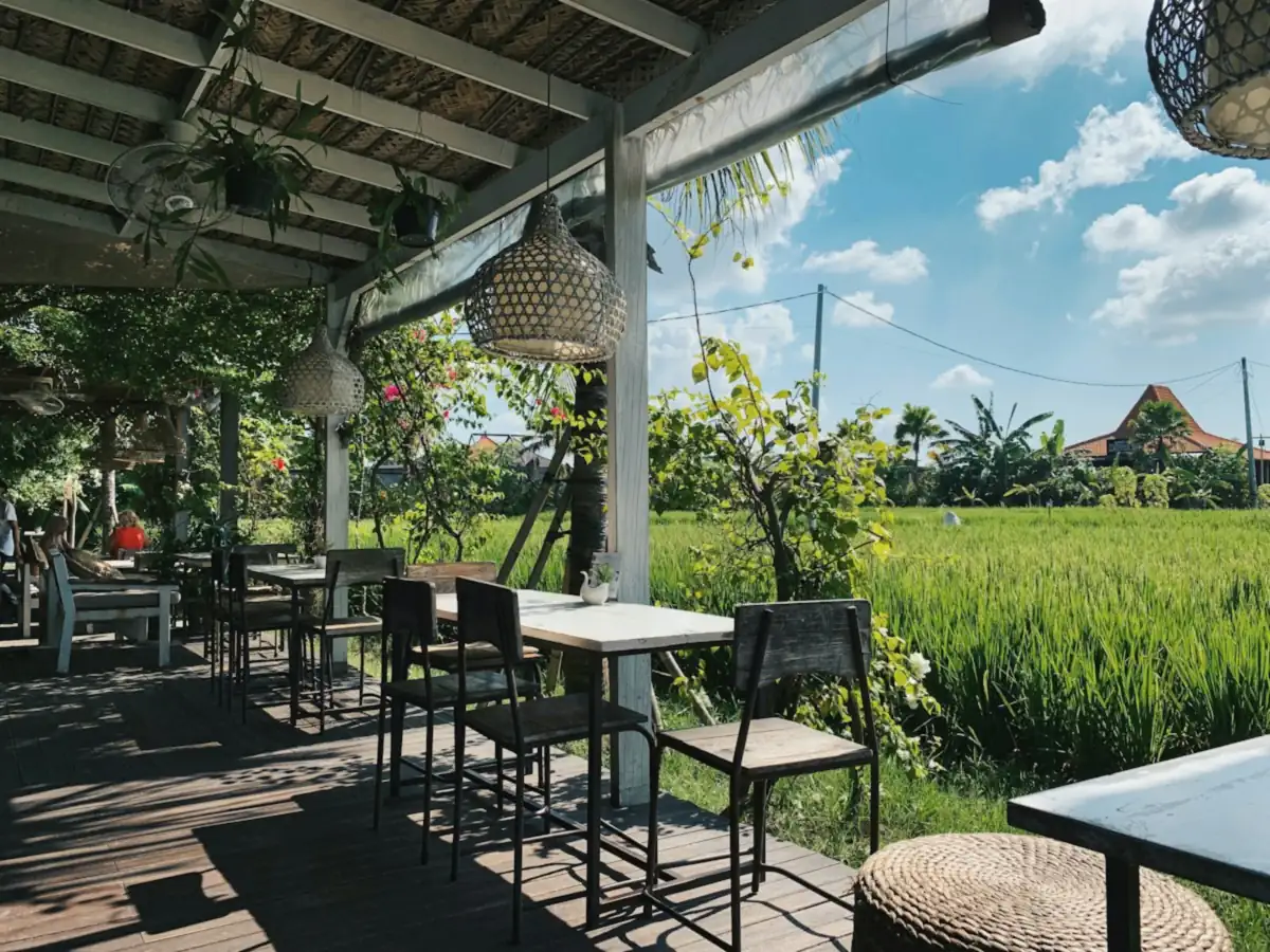 Sunny open-air café terrace with wooden tables, rattan lamps and bougainvillea looking out over green rice fields in Bali