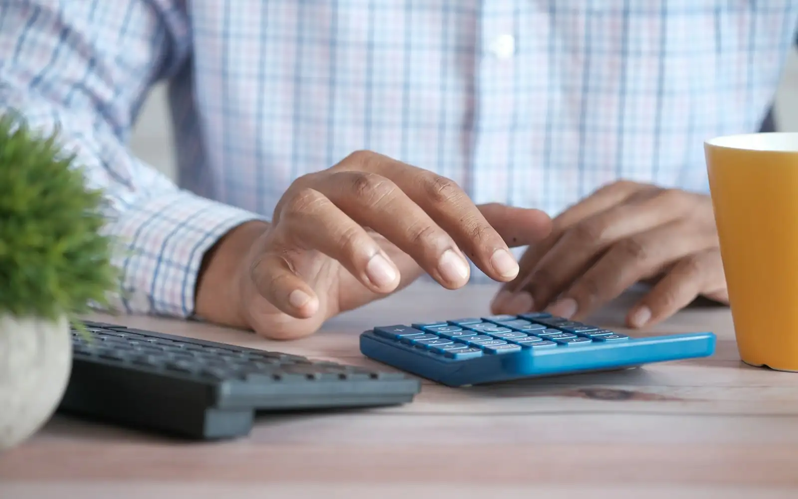 Close-up of hands typing on a blue calculator beside a keyboard, a plant and a coffee mug