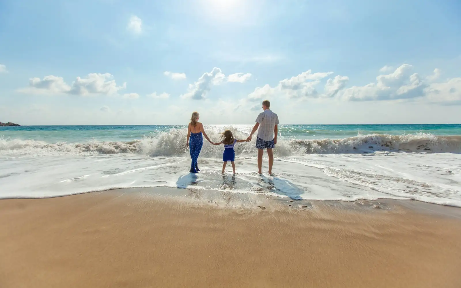 Parents and a young child holding hands as they walk into gentle surf on a sunny beach