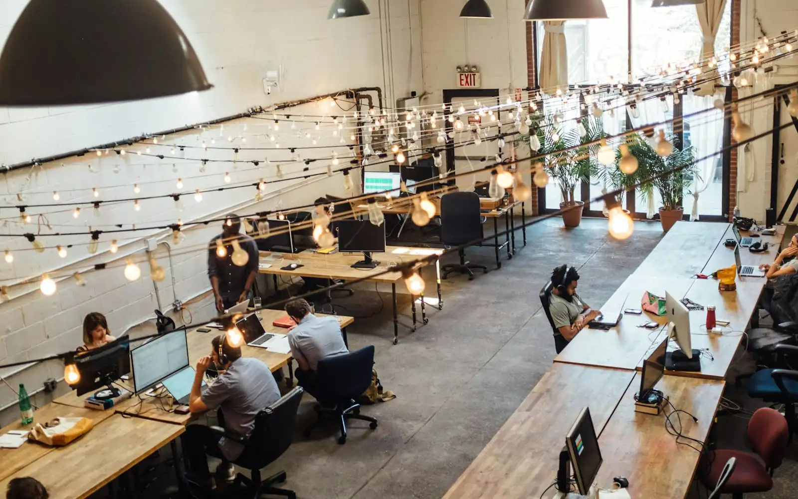 Open-plan coworking space with long wooden desks, people at computers and string lights overhead