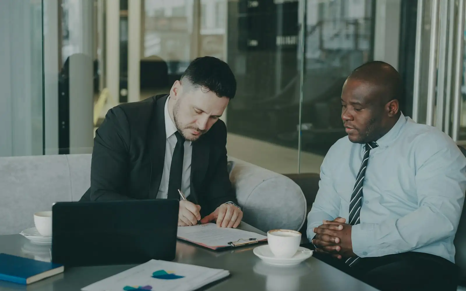 Two businessmen in an office lounge, one signing a document while the other looks on