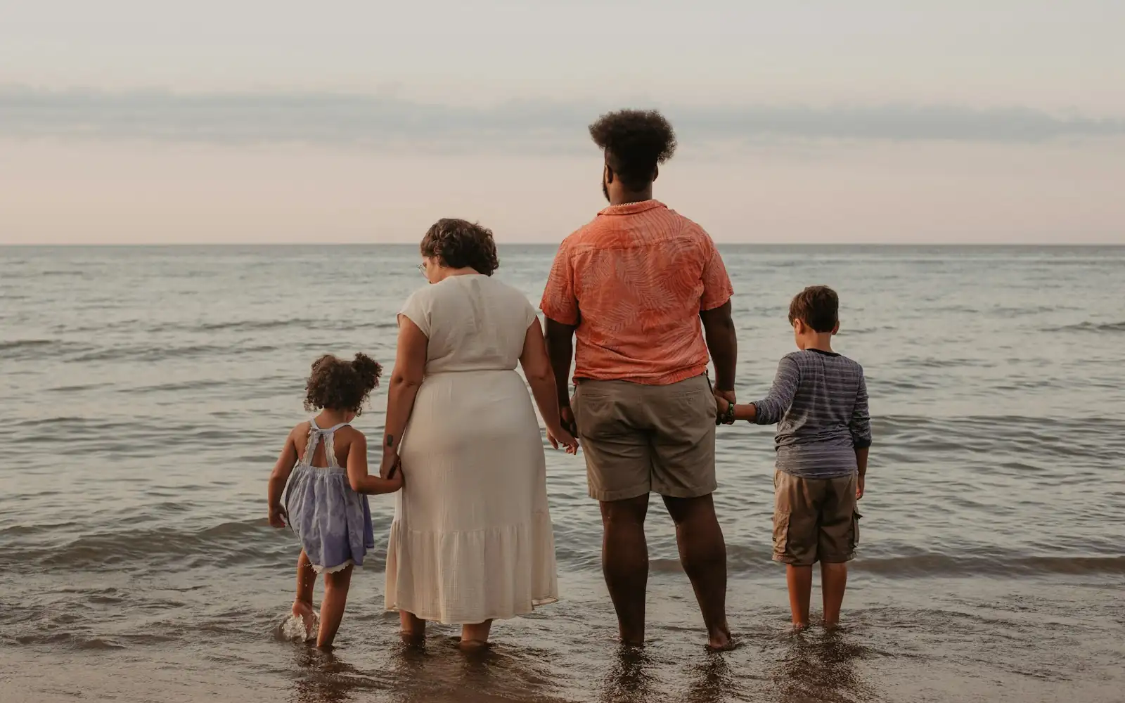 Family of four holding hands while wading in calm shallow sea water in soft evening light