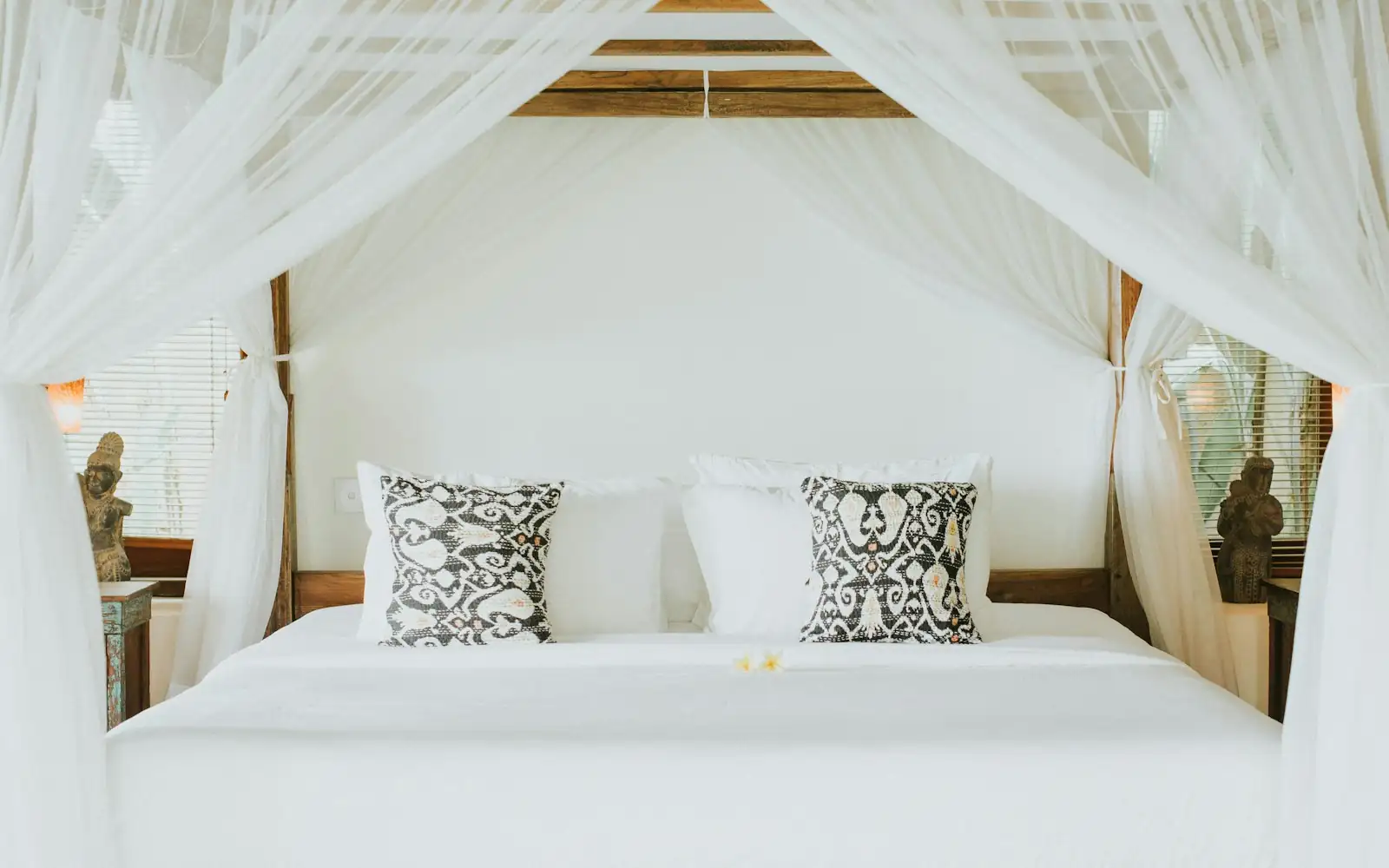 Four-poster bed draped in white mosquito netting with patterned cushions in a calm villa bedroom