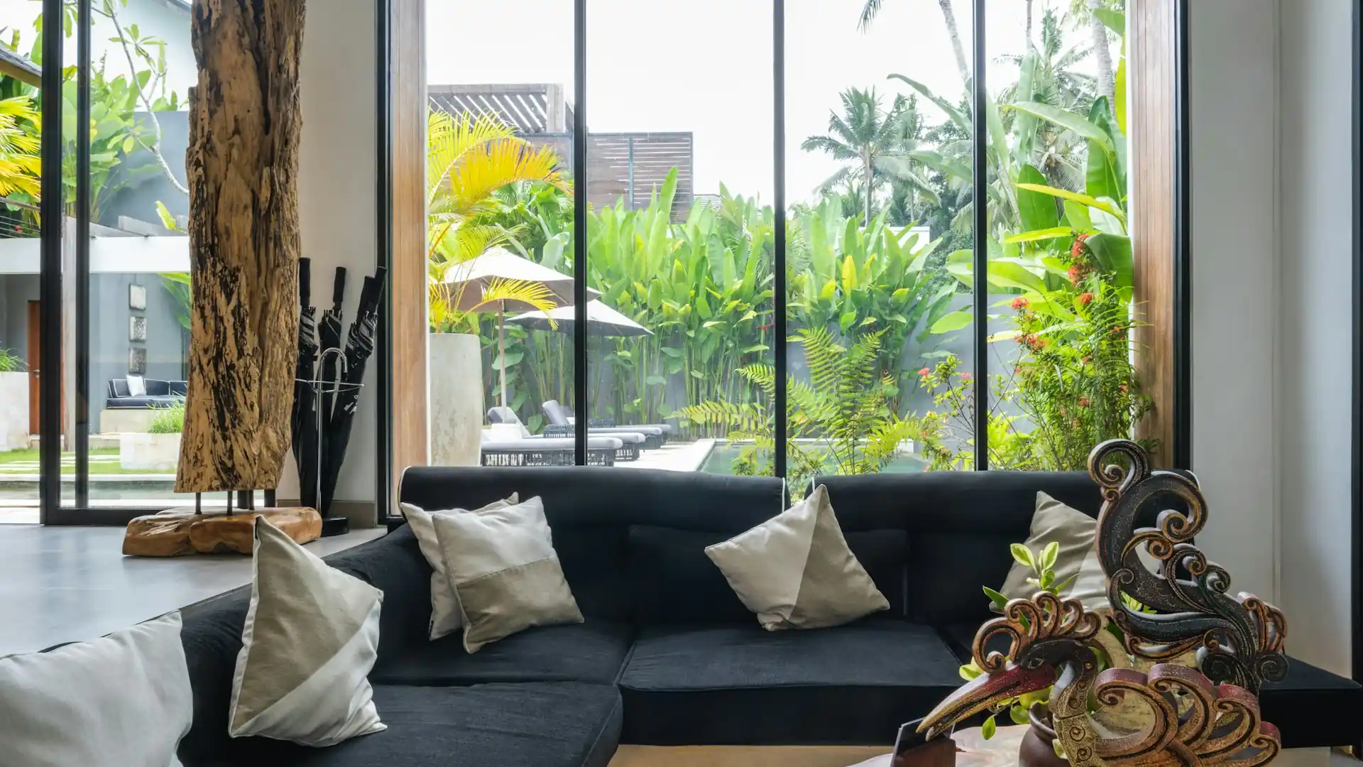 Bright villa living room with a dark sofa and floor-to-ceiling windows opening onto a tropical garden