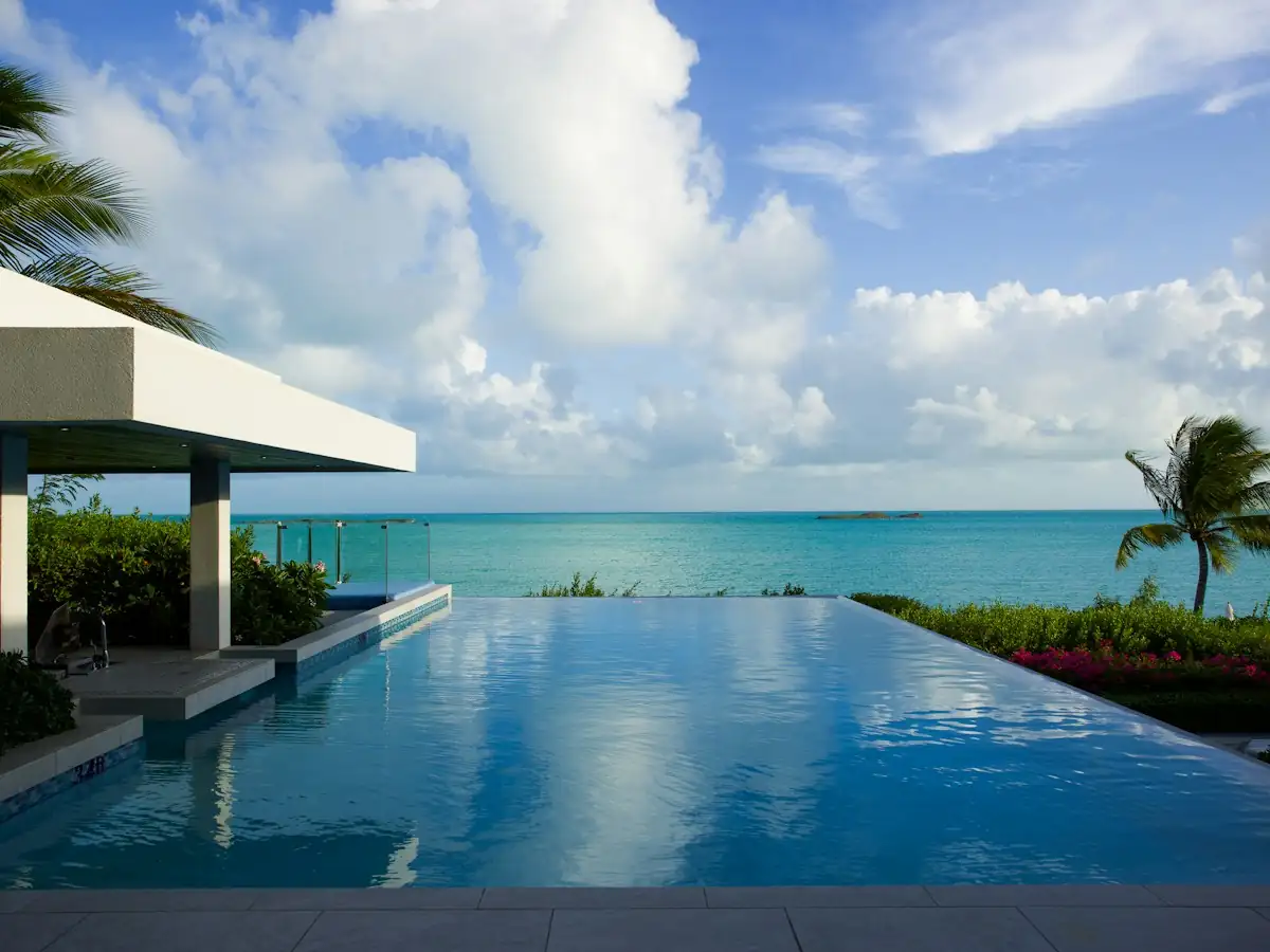 Modern villa infinity pool blending into a turquoise ocean under a bright cloudy sky