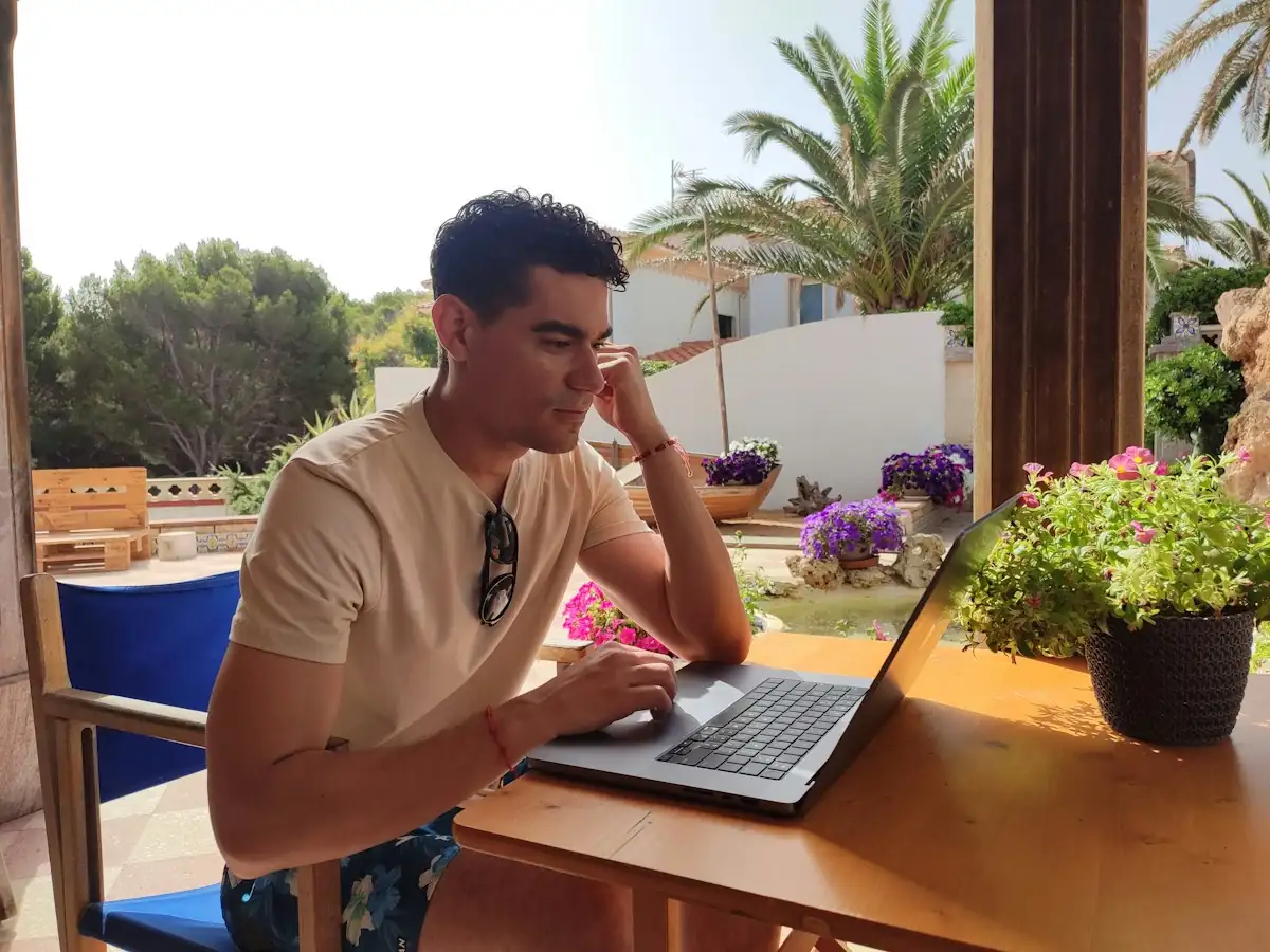 Man working on a laptop at an outdoor table on a sunny garden terrace with flowers and palm trees