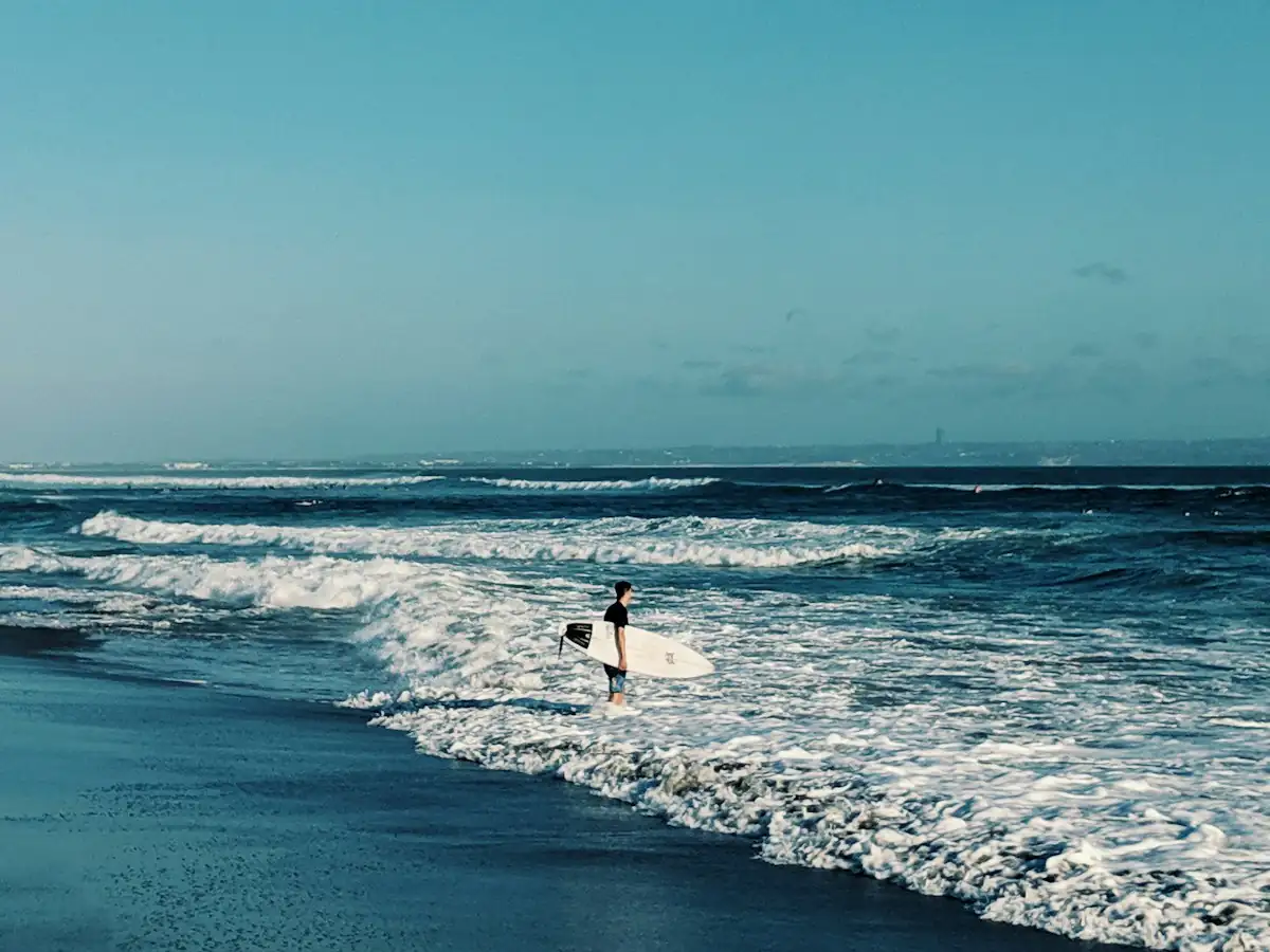 Lone surfer carrying a board into rolling white surf on a dark volcanic sand beach