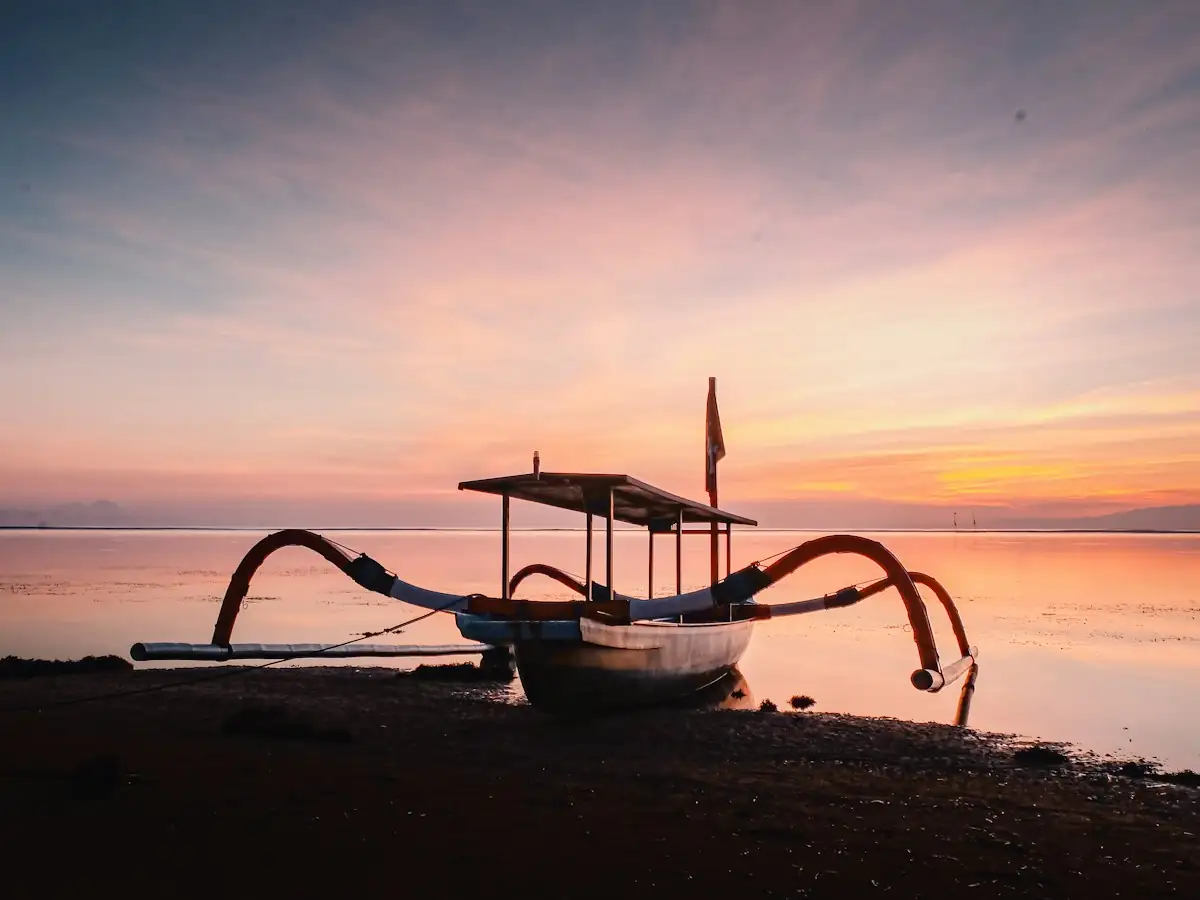 Balinese jukung fishing boat with bamboo outriggers silhouetted against a pink sunrise over calm water