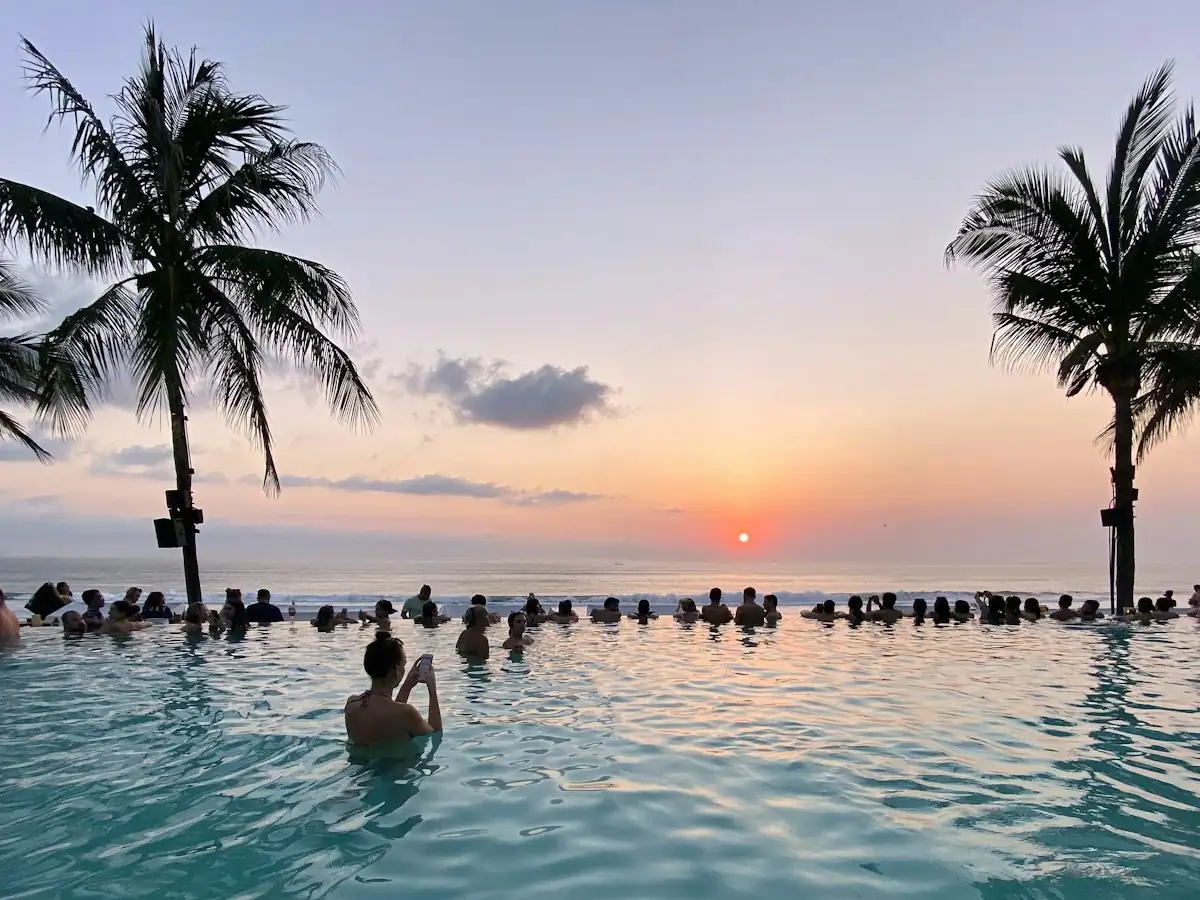 Guests in a beach club infinity pool between coconut palms watching the sun set over the ocean